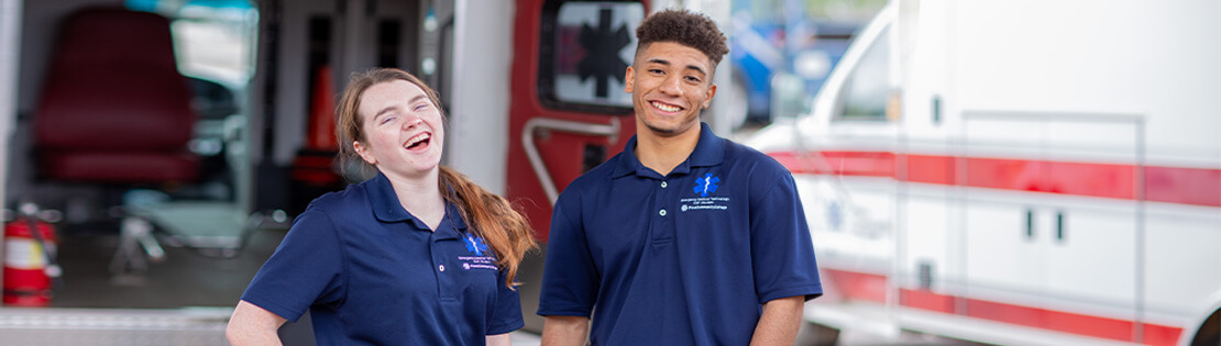 Two paramedic students stand smiling in front of an ambulance