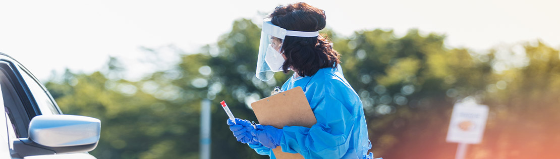 A woman stands in PPE ready to give a COVID test to an upcoming vehicle