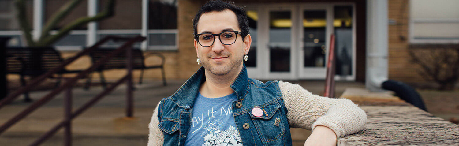 A person stands smiling in a jean jacket in front of a building