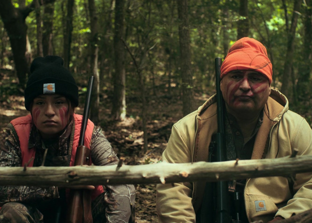 John Proudstar as Leon in 'Reservation Dogs', sits on a log in the woods next to Willie Jack played by Paulina Alexis 