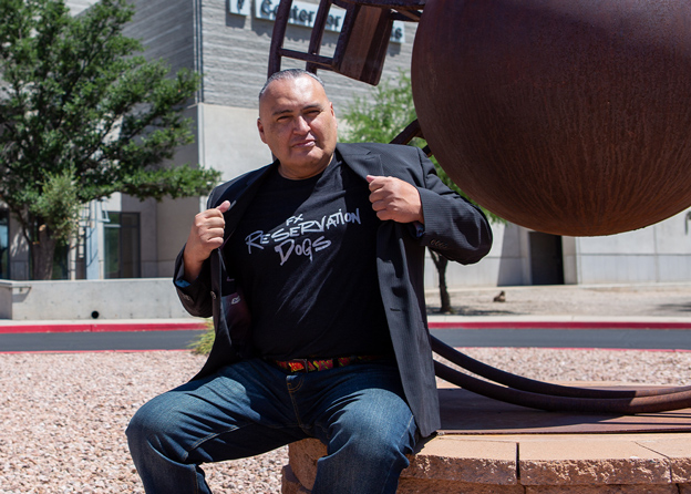 John Proudstar sits in front of the Center of The Arts at PIma West Campus, showing off his Reservation Dogs branded Tshirt