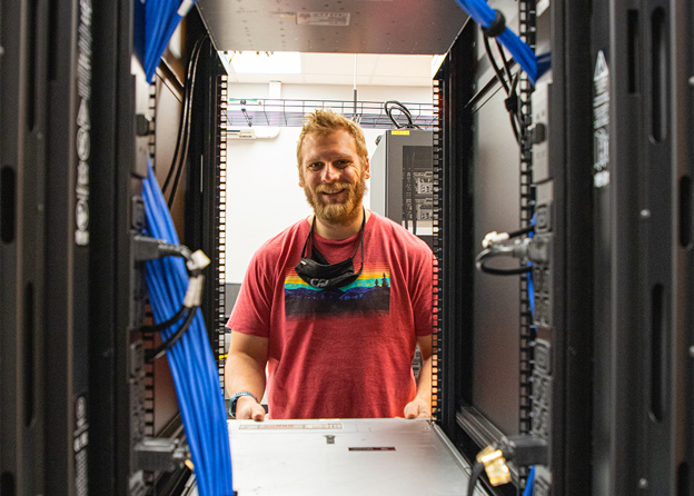 A IT Student stands smiling through a server closet in a server room