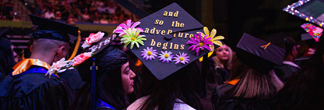 A student sits at graduation, Her mortarboard reads "and so the adventure begins"