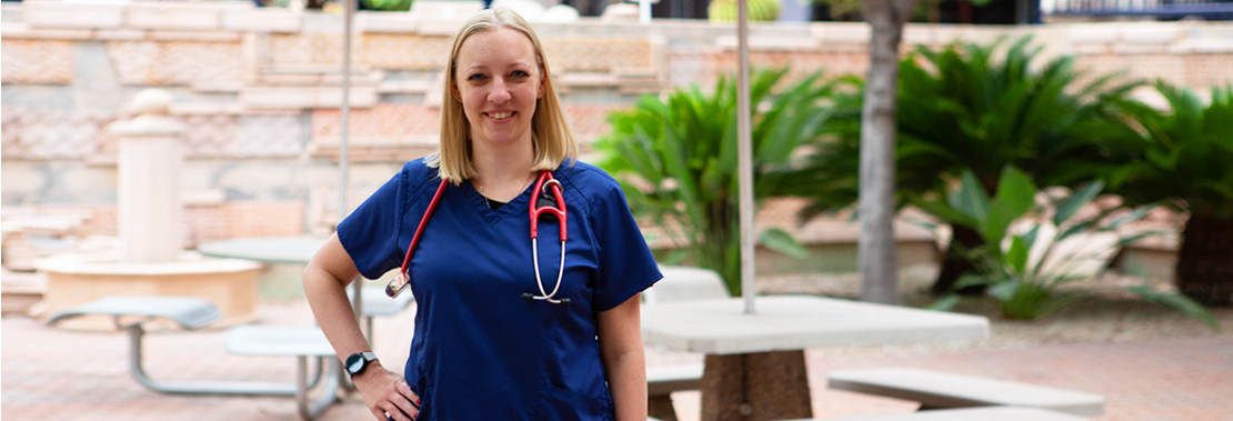 A woman stands in blue nursing scrubs in a courtyard smiling at the camera