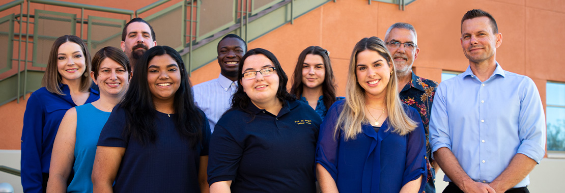 A group of 10 individuals stands smiling in front of a building at Pima's northwest campus.
