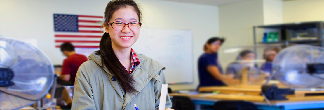 A female student smiles while standing in an engineering classroom