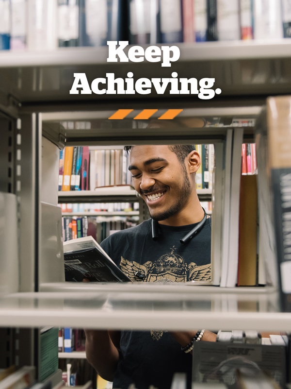 A student is seen through bookshelves reading a book and smiling in a library; Overlay text reads "Keep Achieving."