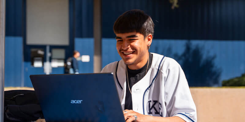 A male student sits working on a laptop in a campus courtyard
