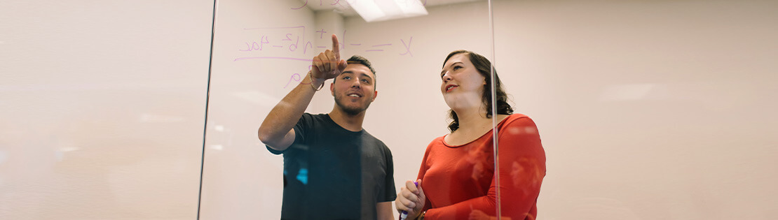 Two Pima students work on a math problem together on a window in a Pima study room
