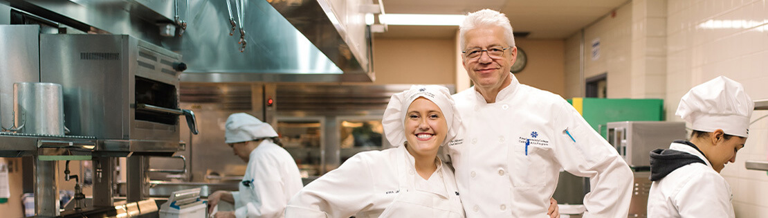 A  Hospitality Fundamentals student and chef stand smiling in a Pima Culinary Kitchen