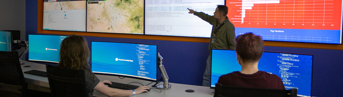 two students sit at computers in Pima's IT Center of Excellence listening to a teacher