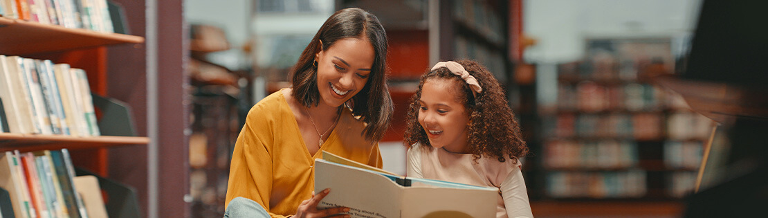 A teacher sits in a library with a young student reading a book