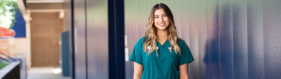 A Student in nursing scrubs stands smiling outside of Pima's West campus