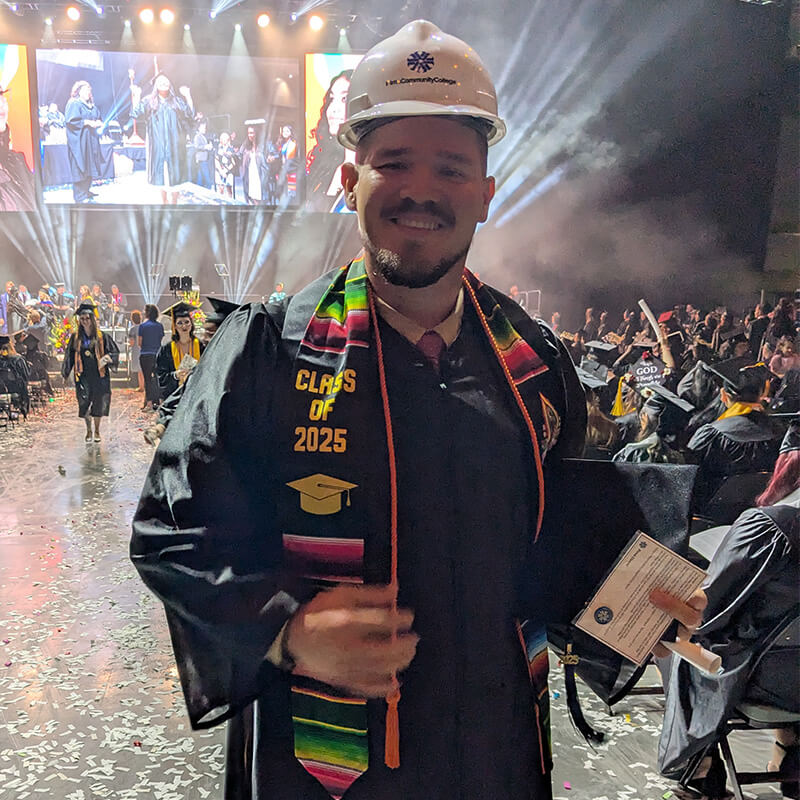 Juan stands at graduation in his graduation gown and hard hat